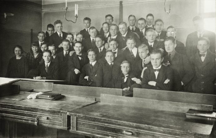 Noin 30 nuorta miestä ja kaksi naista ryhmäkuvassa isossa salissa. Miehillä on mustat puvut ja mirri tai solmio kaulassa.
