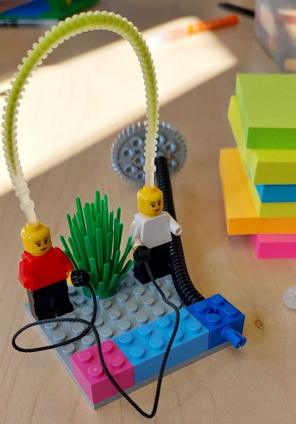 Two LEGO® figurines joined together by two connecting cables, one going from head-to-head, the other going from hand-to-hand. A stack of colorful sticky notes in the back.