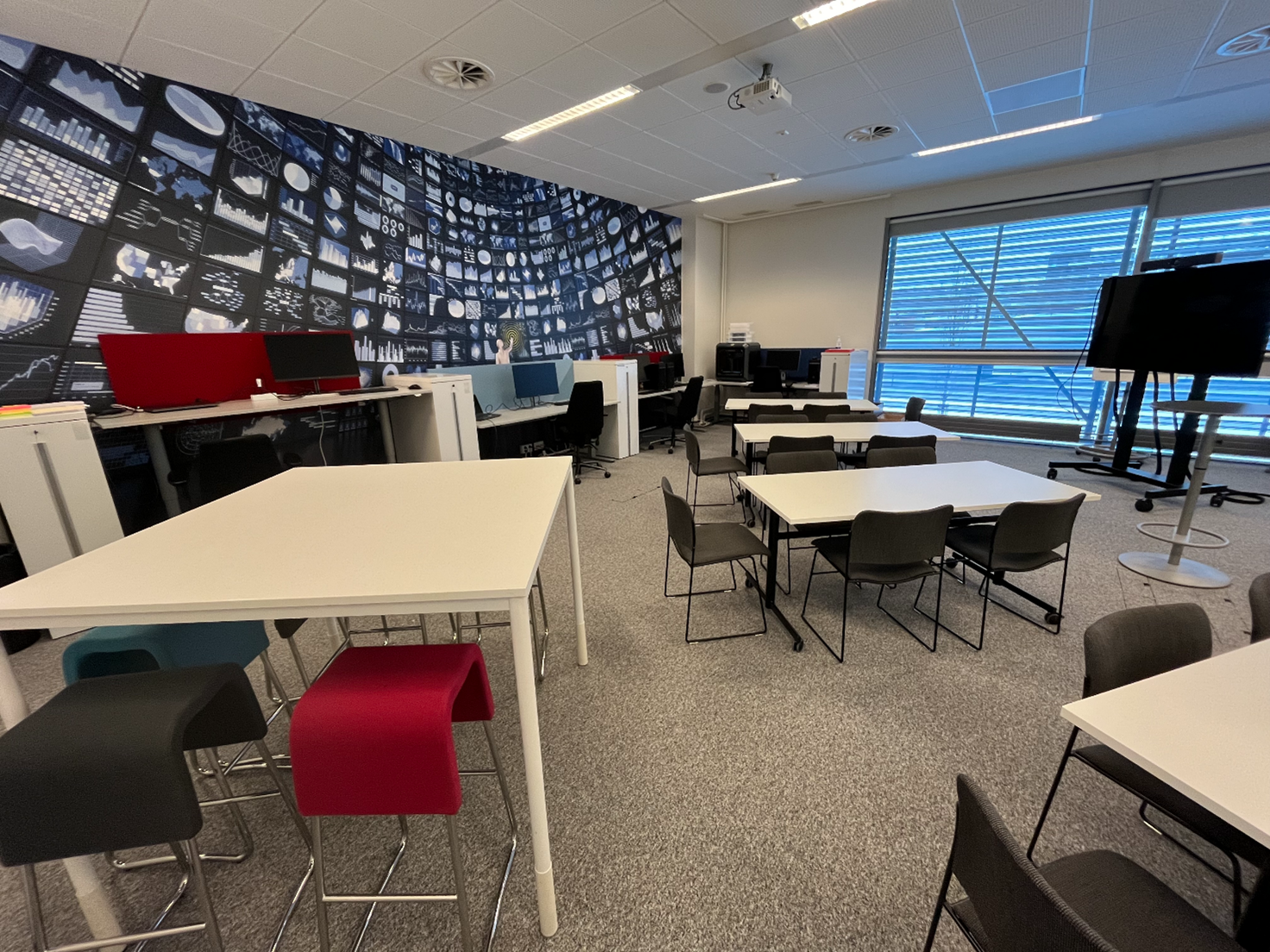 Picture of the actual living lab space with chairs and tables and workstation desks as well as a monitor and a 3D printer.