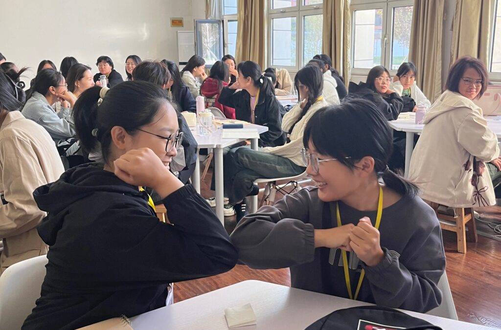 Kaksi kiinalaista opiskelijaa hymyilee pöydän ääressä toisilleen, kyynärpäät yhdessä kuin toisiaan tervehtien. Taustalla näkyvät heidän luokkatoverinsa työn touhussa.