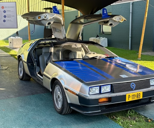 A silver DeLorean DMC-12 sports car with its distinctive gull-wing doors open is parked outdoors.