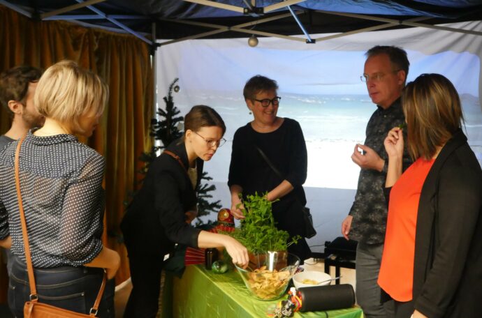 Kuvassa henkilöitä keskustelemassa ja maistelemassa pöydänantimia moniaistisessa tilassa. 