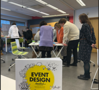 : Kuvassa näkyy koulutukseen osallistuneita henkilöitä, jotka ovat kerääntyneet pienryhmässä pöydän ympärille. Lisäksi kuvassa näkyy kirja nimeltä Event Design, johon Tapahtumamuotoilun koulutus perustuu.