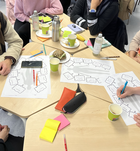 Six people around a table with sticky notes, felt pens, templates showing personas, coffee mugs. bread rolls, water bottles and other clutter.