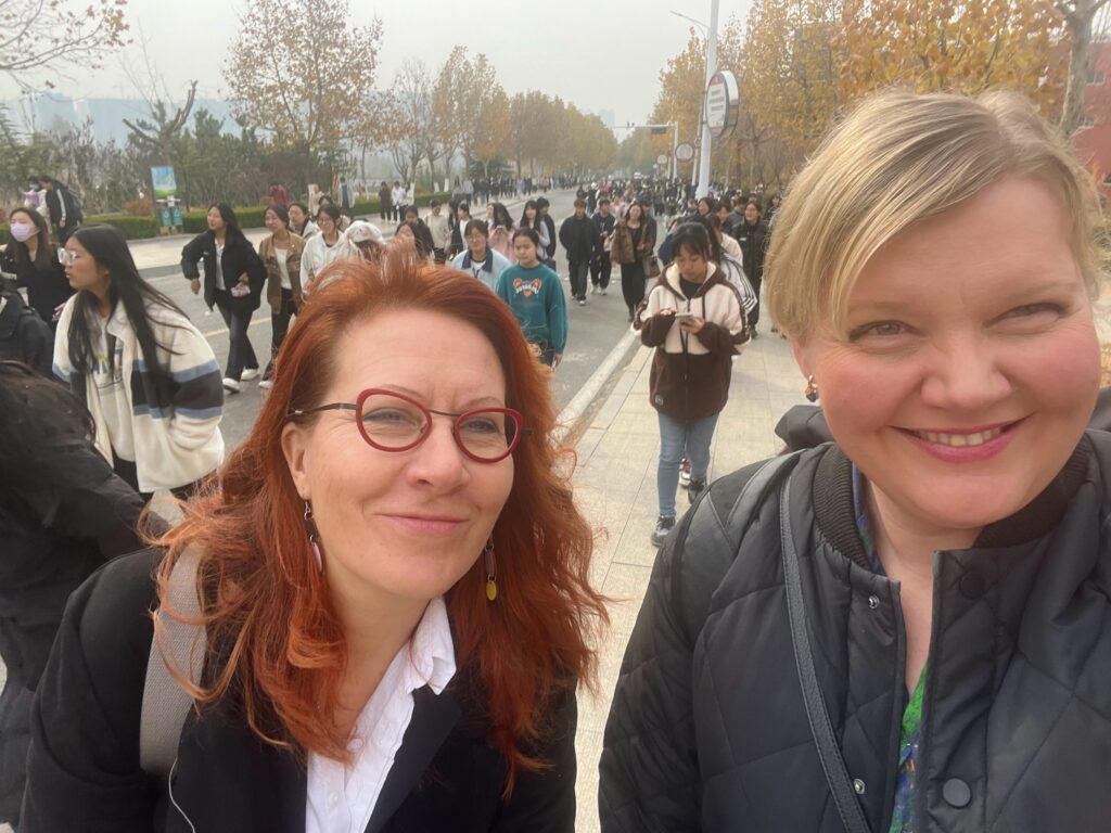Selfiekuvassa on kaksi Laurean opettajaa hymyilemässä kampuksella tunneille taivaltavan opiskelijavirran keskellä.