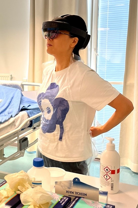 ALT-teksti: Sairaanhoitajaopiskelija katselee virtuaalista ohjetta suojainten pukemisesta HoloLens 2- älylaseilla ja toimii samanaikaisesti ohjeiden mukaisesti pukien esiliinaa.